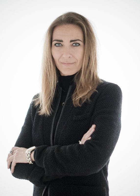 En kvinde med langt, lysebrunt hår står selvsikkert med armene over kors, iført en sort strukturjakke og sølvarmbånd, på en hvid baggrund - og præsenterer en privat socialrådgivers afklarede look.
