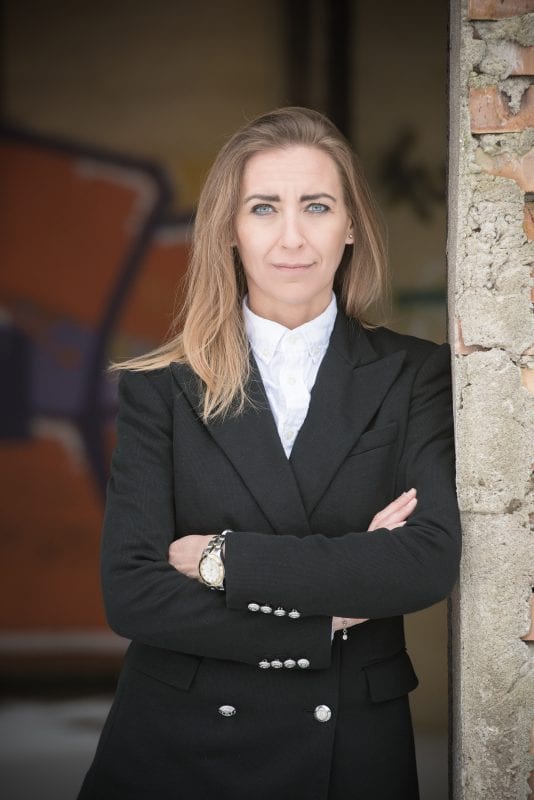 En privat socialrådgiver med langt blondt hår, iført sort blazer og hvid skjorte, står med armene over kors op ad en vejrbidt mur. Hun kigger selvsikkert ind i kameraet, mens graffiti dekorerer den slørede baggrund.