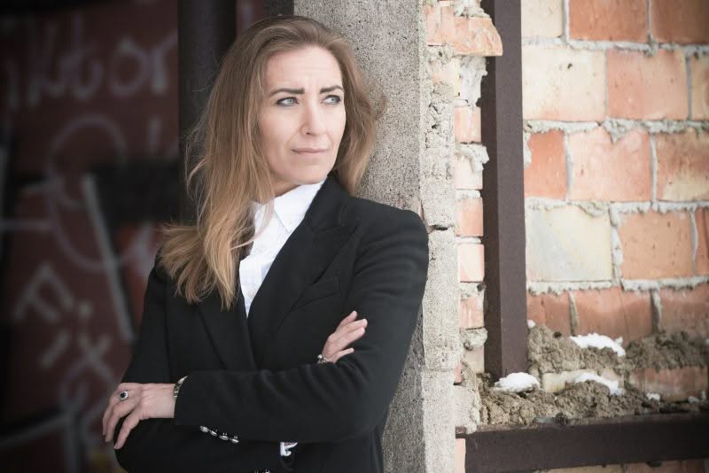En privat socialrådgiver i sort blazer står med armene over kors og læner sig op ad en betonsøjle. Hun kigger til siden ved siden af en murstensvæg med pletter af sne og blottet mørtel.