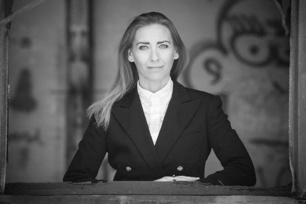En privat socialrådgiver med langt hår, klædt i mørk blazer og hvid skjorte, står selvsikkert ved en vinduesramme og kigger fremad. Det sort-hvide foto har sløret graffiti i baggrunden.