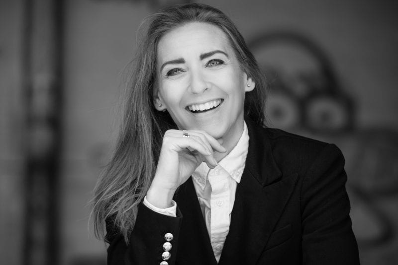 Sort-hvidt portræt af en smilende privat socialrådgiver i tilknappet blazer og hvid skjorte, der hviler hagen på sin hånd, med langt hår over den ene skulder og en sløret baggrund.