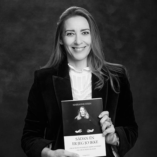 En kvinde med langt hår, iført mørk blazer og lys bluse, smiler, mens hun holder bogen SÅDAN ÉN ER JEG JO IKKE af privat socialrådgiver Marianne Stein. Baggrunden er mørk og ensfarvet. Billedet er i sort og hvid.