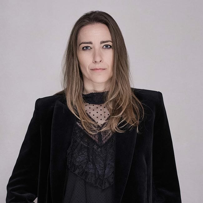 En privat socialrådgiver med langt, glat brunt hår i sort fløjlsblazer og blondetop står på en almindelig lysegrå baggrund og ser direkte ind i kameraet med et neutralt udtryk.