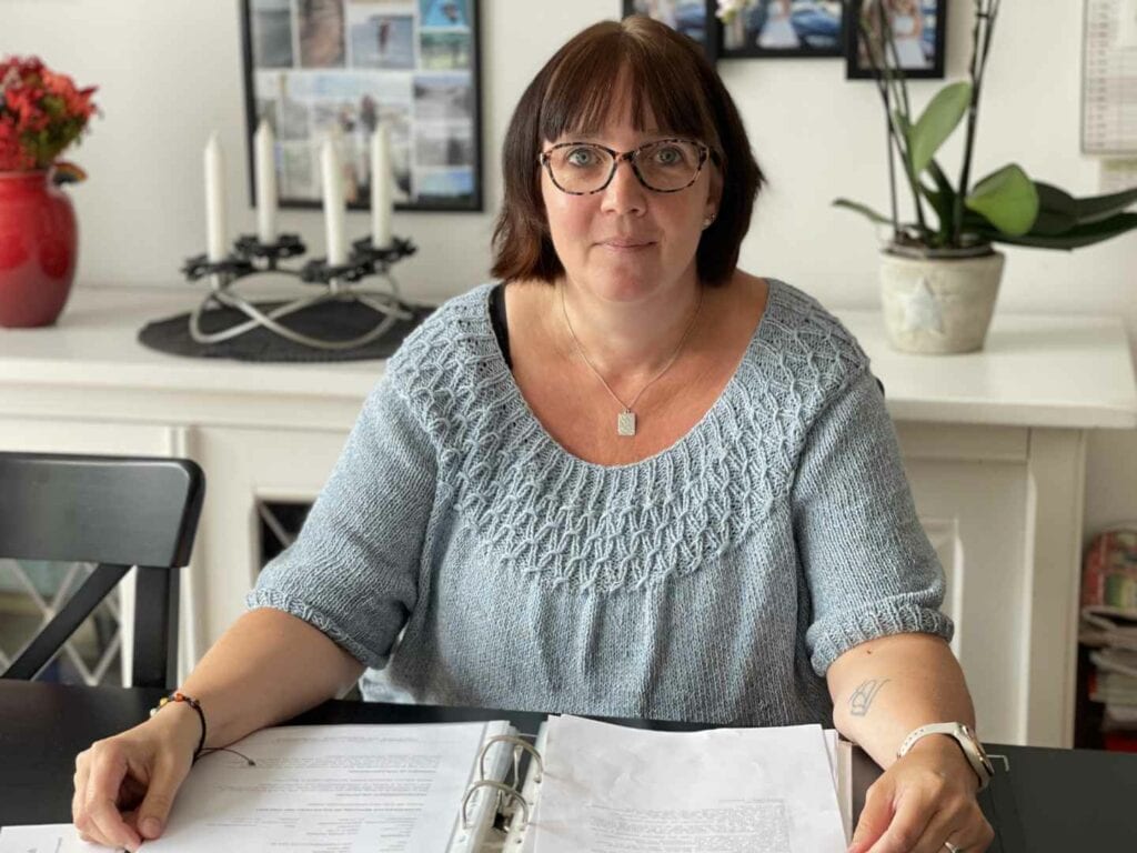 En privat socialrådgiver med briller og en lyseblå strikket top sidder ved et bord med et ringbind og papirer. Bag hende står indrammede fotos, en orkidé i potte, en lysestage og en rød vase med blomster.