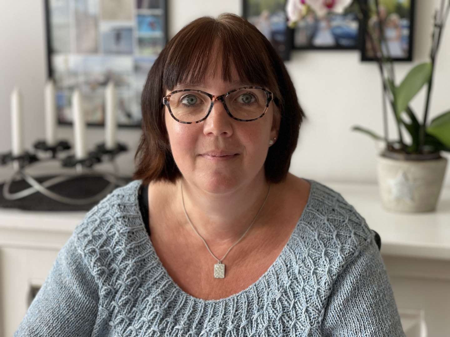 En kvinde med kort brunt hår og briller, iført en lyseblå striktrøje og en sølvhalskæde, sidder indendørs som privat socialrådgiver foran en hvid baggrund med en plante og indrammede billeder.